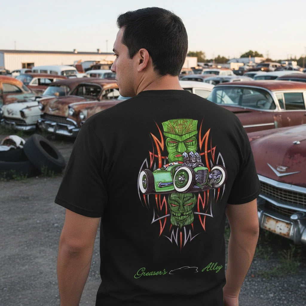 A man in the black Greaser's Alley Rod and Tiki tee, walking through a yard of 1950s cars at sunset