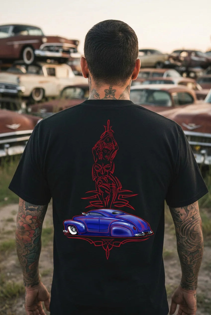 Back view of a tattooed man in a black Greaser's Alley tee with a red pinstripe pin-up and blue custom lead sled print, standing in a field of stacked classic cars at dusk